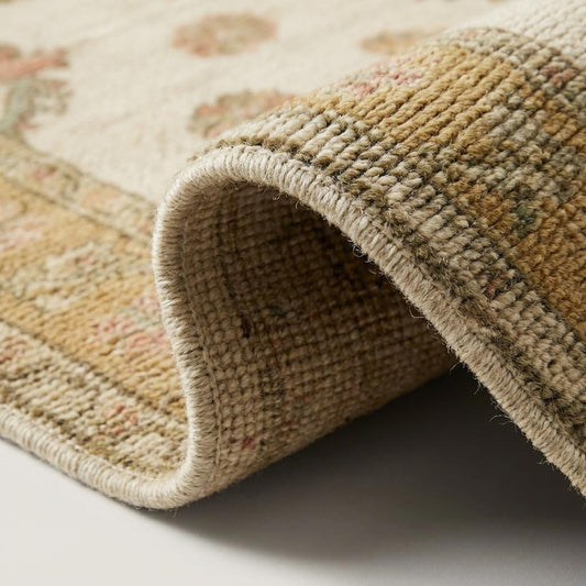 Close-up of hand knotted wool rug showing individual knot definition, dense wool pile, ivory base, and muted gold floral detailing with visible artisan weave structure.