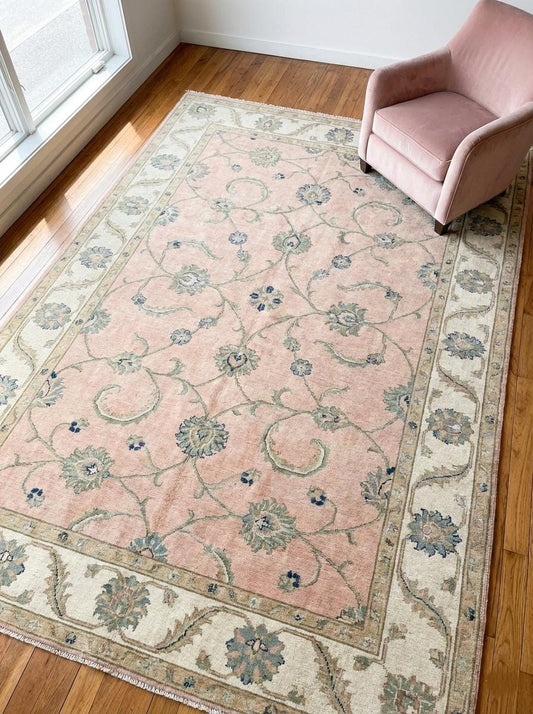 Hand knotted Oushak rug with blush pink background and cream floral border on wood floor.