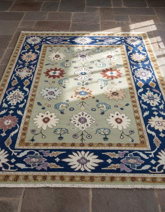 Top view of sage green floral area rug with traditional Oushak design and navy border on stone floor