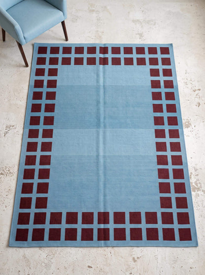Handwoven sky blue flatweave dhurrie rug with maroon square border in living room setting