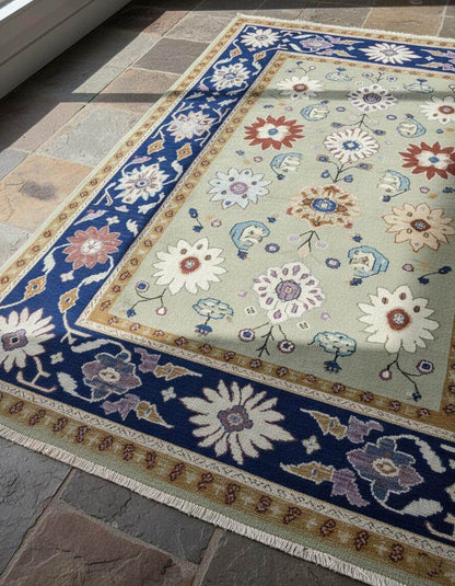 Top view of sage green floral area rug with traditional Oushak design and navy border on stone floor